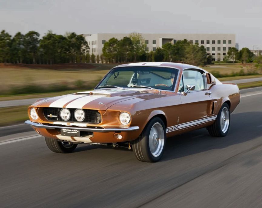 Front three-quarter rolling action shot of a 1967 Revology Shelby GT350 in Bronze Metallic with white Le Mans stripes driving on a highway.
