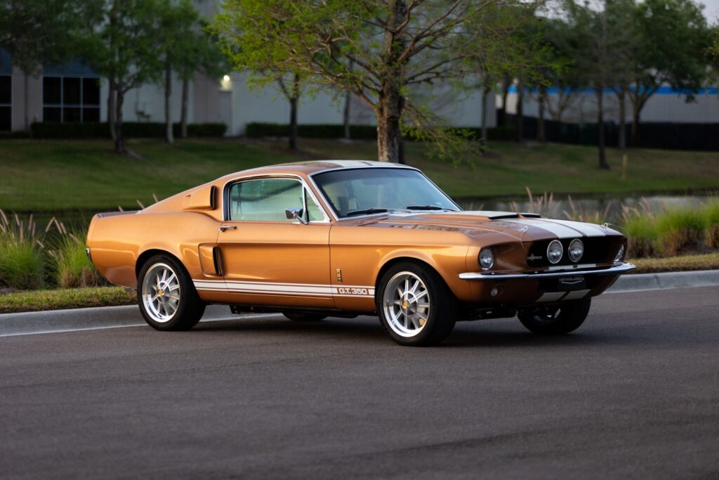 Front three-quarter view of a Copper metallic 1968 Revology Shelby GT350 Fastback with white Le Mans stripes, parked on a suburban road at dusk.