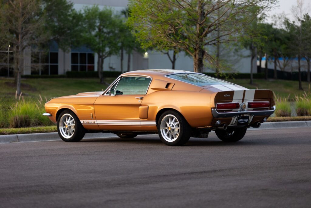 Rear three-quarter view of a Copper metallic 1967 Revology Shelby GT350 Fastback with white racing stripes, showcasing the horizontal taillights and 10-spoke aluminum wheels.