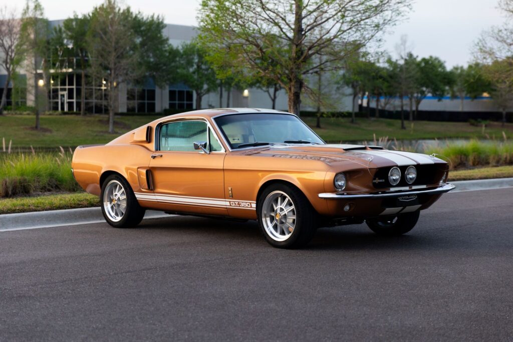 Low-angle front three-quarter view of a Copper metallic 1967 Revology Shelby GT350 with center-mounted grill lights and white Le Mans racing stripes.