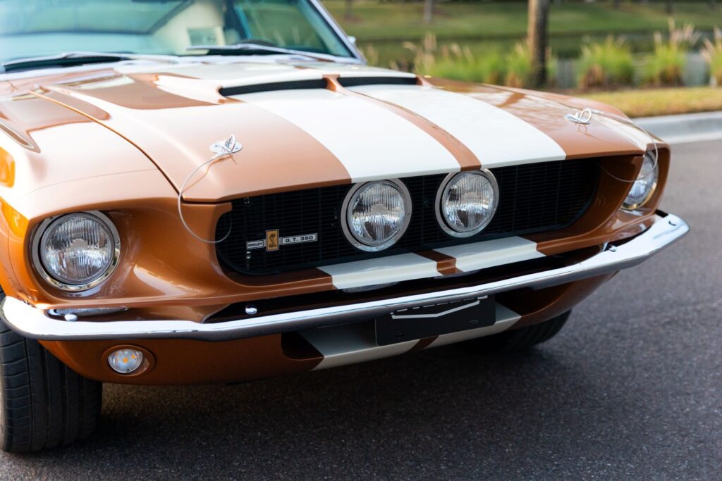 Close-up of the front grill of a Copper metallic 1967 Revology Shelby GT350, featuring center-mounted driving lights, white racing stripes, and chrome hood pins.