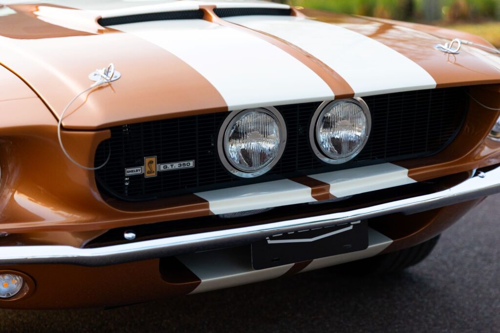 Extreme macro of a 1967 Revology Shelby GT350 front grill in copper metallic, showing center-mount high-beam lights, chrome hood pins, and white Le Mans stripes.