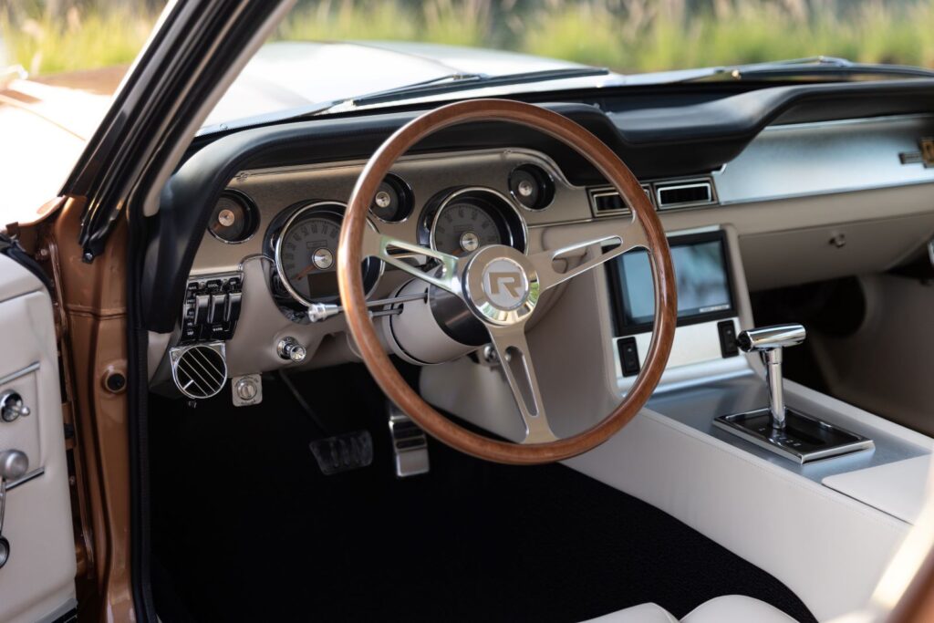 Driver-side cockpit view of a 1967 Revology Shelby GT350 featuring a wood-rimmed steering wheel, modern digital gauges, and an integrated touchscreen infotainment system.