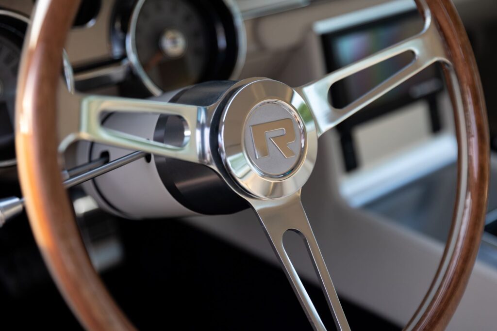 Close-up of the polished aluminum three-spoke wood-rimmed steering wheel with the Revology "R" logo center cap on a 1967 Shelby GT350.