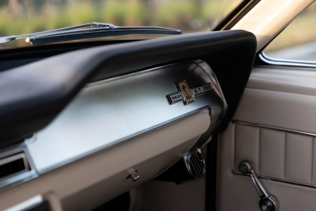 Close-up of the brushed aluminum passenger dashboard inlay and "Shelby G.T. 350" emblem on an off-white leather 1967 Revology Shelby.