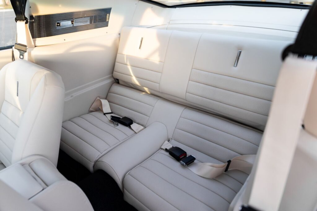 Rear cabin view of a 1967 Revology Shelby GT350 with off-white leather fold-down rear seats and modern three-point seatbelts.