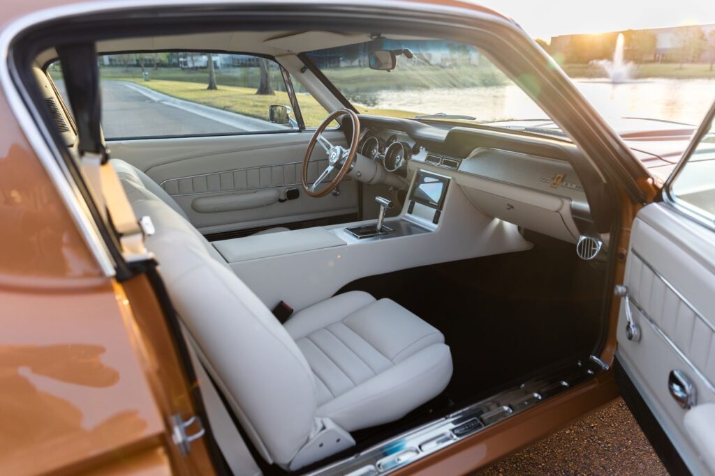 Wide-angle view from the passenger door of a 1967 Revology Shelby GT350 interior with off-white leather sport seats, a custom center console, and a wood-rimmed steering wheel.