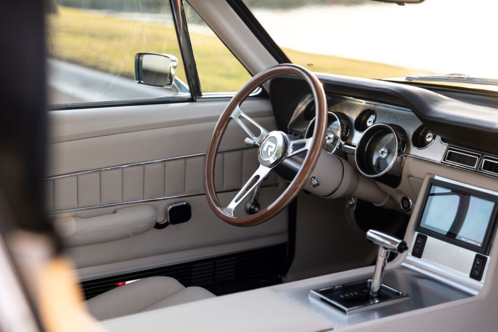 Interior view of the 1967 Revology Shelby GT350 cockpit, featuring a touchscreen infotainment system, polished T-handle automatic shifter, and a wood-rimmed steering wheel with the Revology "R" logo.