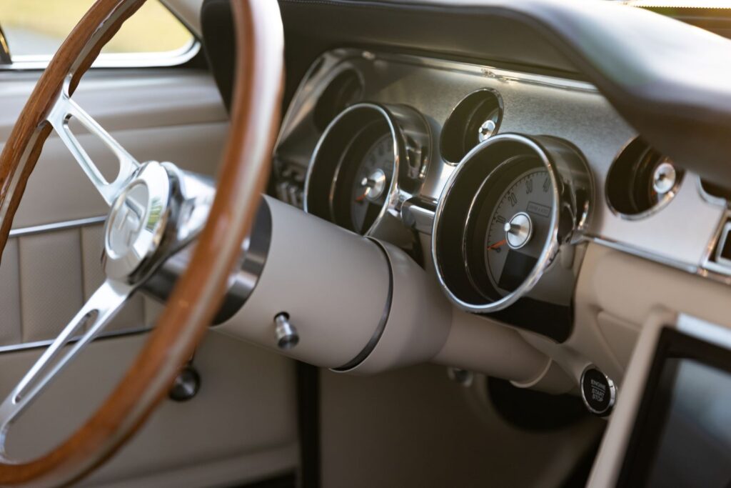 Side-profile close-up of the steering column, wood-rimmed steering wheel, and modern electronic gauges in a 1967 Revology Shelby GT350 with brushed aluminum dash trim.