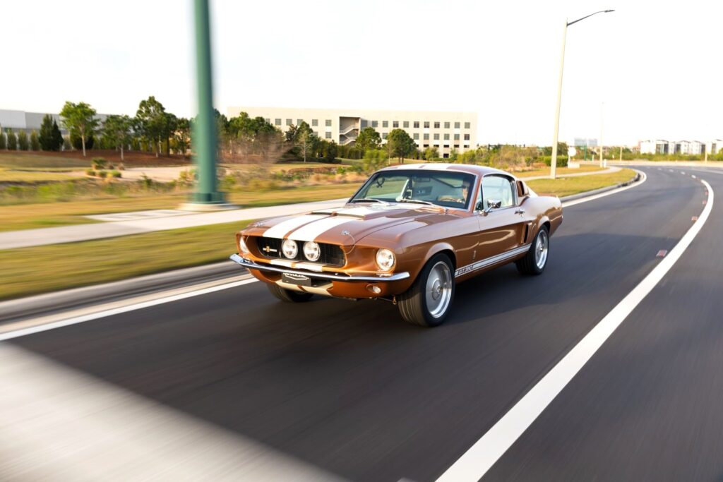 Action shot of a Copper metallic 1967 Revology Shelby GT350 with white racing stripes driving on an open highway at sunset.