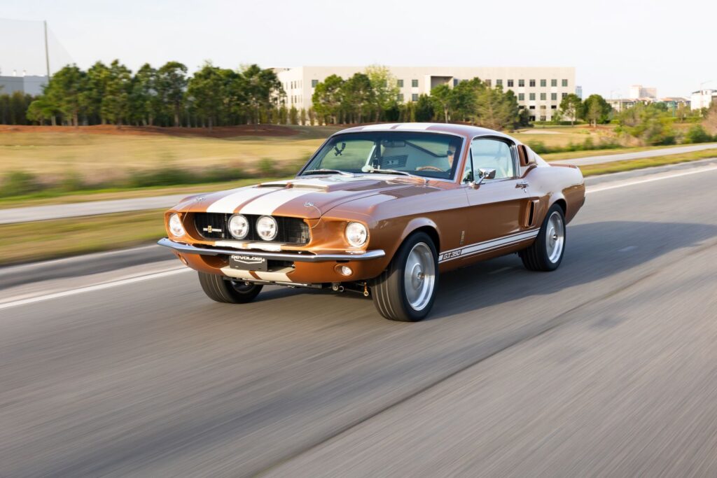 Front three-quarter action shot of a Copper metallic 1967 Revology Shelby GT350 with white Le Mans stripes driving on a paved road.