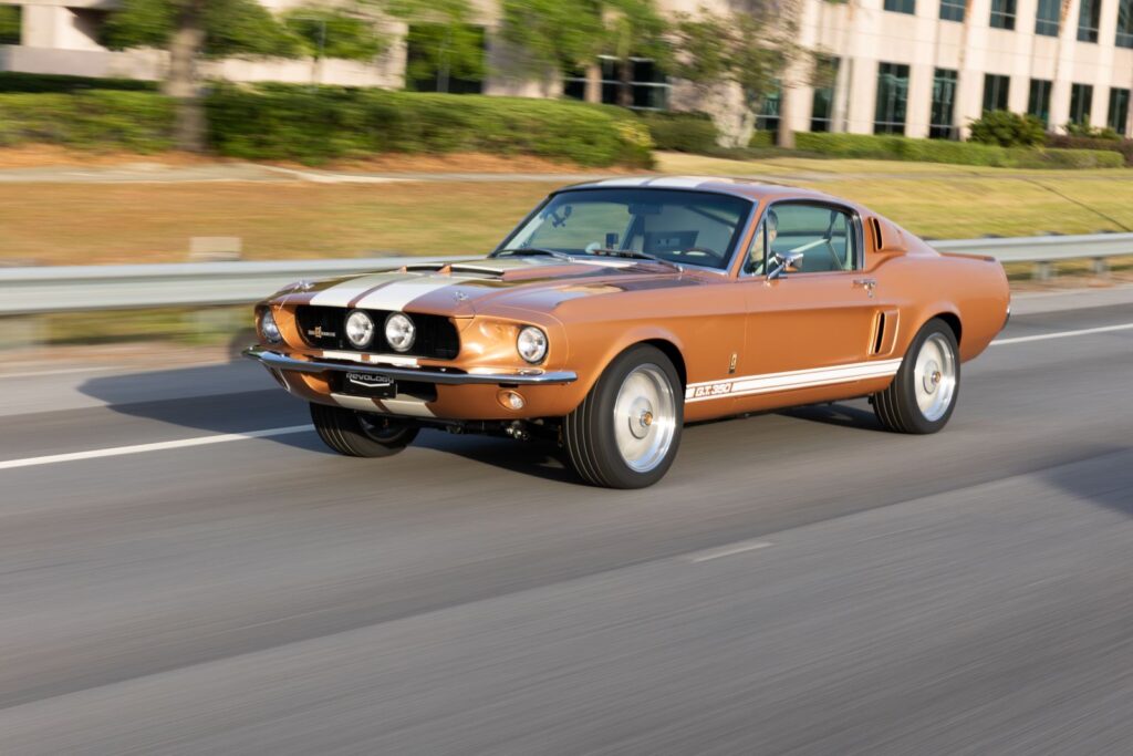 Side-angle rolling action photograph of a Copper metallic 1967 Revology Shelby GT350 with white Le Mans stripes driving at highway speeds.