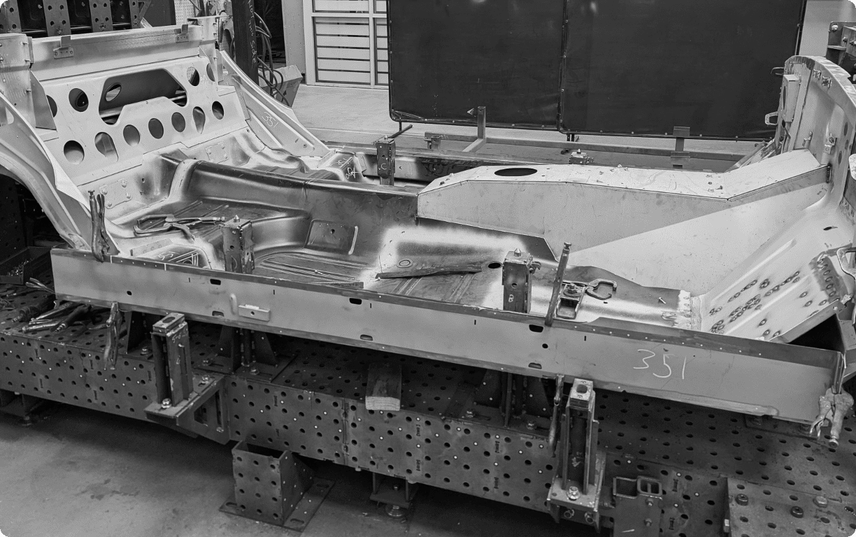 B&W documentary-style photo of a raw, bare-metal classic Mustang body structure during production at Revology Cars. The complex perforated framing fixture holds the rocker panel, floor pan, and bulkhead for precise alignment. Metal fabrication tools and handwritten numbers like "351" are visible on the raw steel.