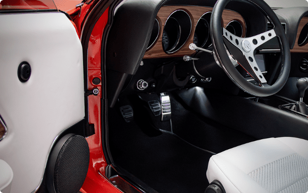 Interior footwell of a Revology 1969 Mustang Boss 429 showing custom billet aluminum performance pedals, parchment white leather seats, and a black leather-wrapped dashboard with walnut wood grain.