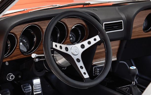 Interior view of a Revology 1969 Mustang Boss restomod featuring a leather-wrapped three-spoke steering wheel with "R" logo, walnut wood grain dashboard, and modern manual shifter.