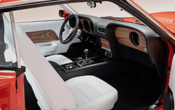 Interior of a Revology 1969 Ford Mustang Boss 429 featuring white leather high-back bucket seats, walnut wood grain dash inserts, and a black leather-wrapped center console with a manual shifter.