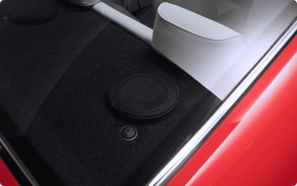 Close-up view through the rear window of a Revology classic Mustang restomod, showcasing the rear package shelf with integrated Harmon-Kardon audio components, including a large black speaker grille and smaller tweeter, set into black carpeting next to a white leather panel.