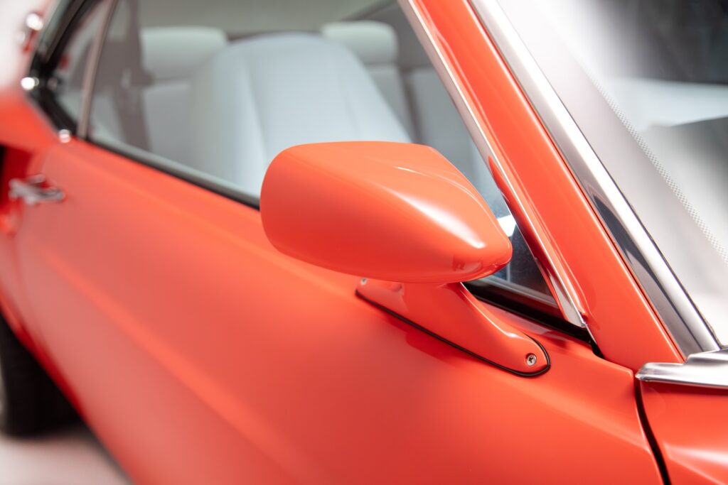 Close-up of the driver-side aerodynamic power mirror on an orange Revology 1969 Ford Mustang Boss 429, featuring matching body color paint and a glimpse of the white leather interior.