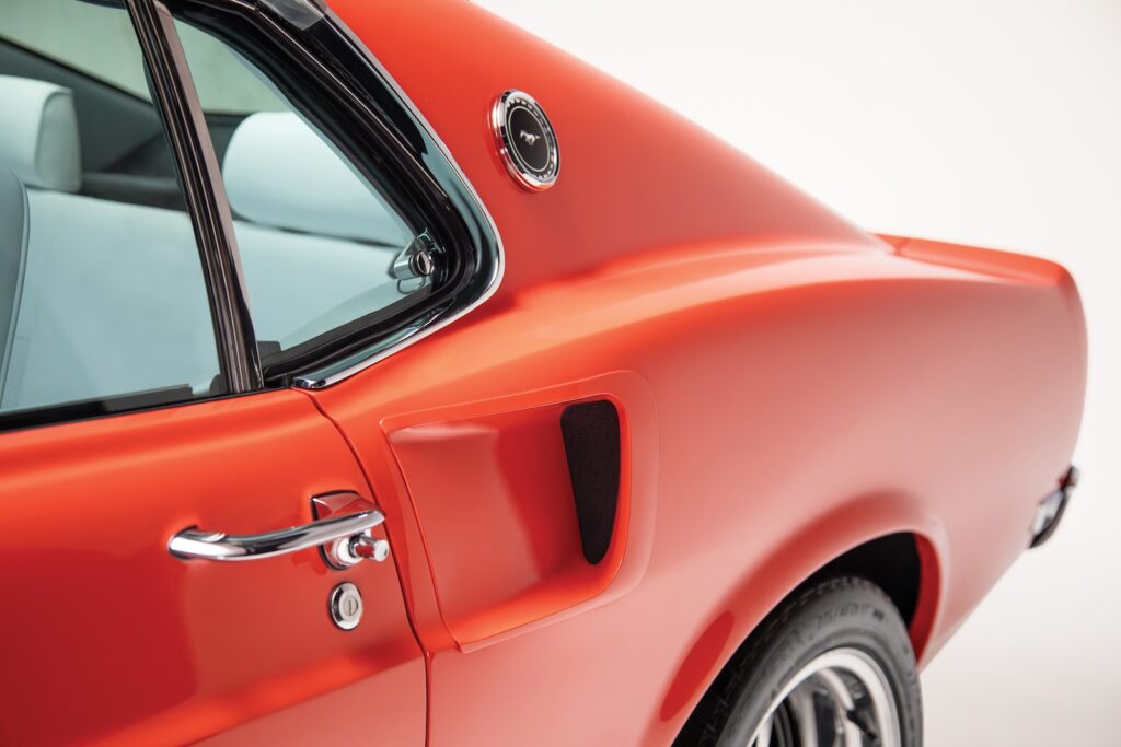 Close-up of the rear quarter panel and C-pillar on an orange Revology 1969 Mustang Boss 429, featuring a functional side scoop, chrome door handle, Mustang C-pillar emblem, and a glimpse of the parchment white leather interior.