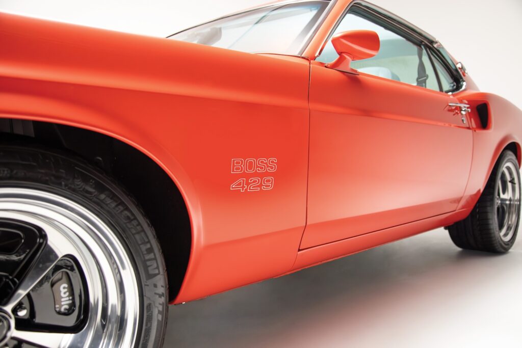 Low-angle view of the front driver-side fender on an orange Revology 1969 Mustang Boss 429, featuring "BOSS 429" decals, Wilwood high-performance brakes, and Michelin Pilot Sport tires.
