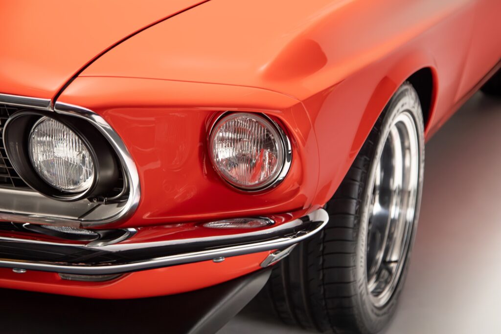 Close-up of the front driver-side headlight, chrome bezel, and bumper of a Revology 1969 Mustang Boss 429 in high-gloss orange, showing the precise fitment and Michelin performance tire.