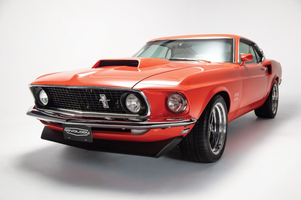 Front three-quarter studio view of a Revology 1969 Ford Mustang Boss 429 restomod in high-gloss orange, featuring the iconic Boss hood scoop, front chin spoiler, and modernized Magnum 500 wheels.
