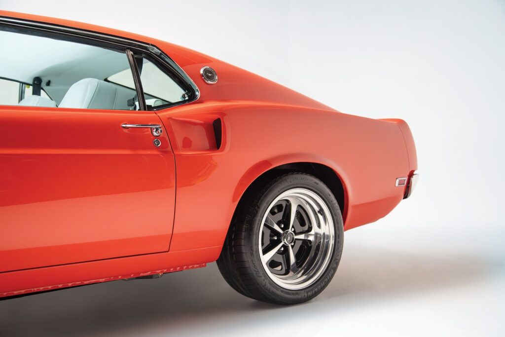 Rear quarter view of an orange Revology 1969 Ford Mustang Boss 429 restomod, highlighting the functional side scoop, C-pillar emblem, 17-inch Magnum 500 wheels, and parchment white leather interior.