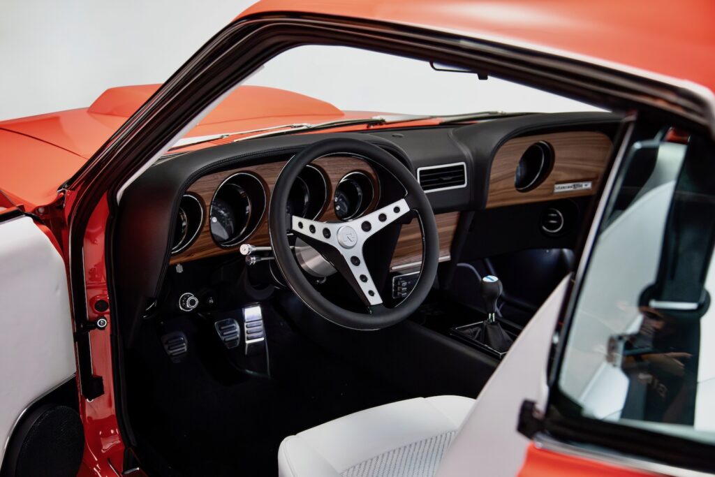Driver's side cockpit view of a Revology 1969 Ford Mustang Boss 429, featuring a black leather-wrapped dashboard with walnut wood grain, a three-spoke aluminum steering wheel, billet pedals, and parchment white leather seats.