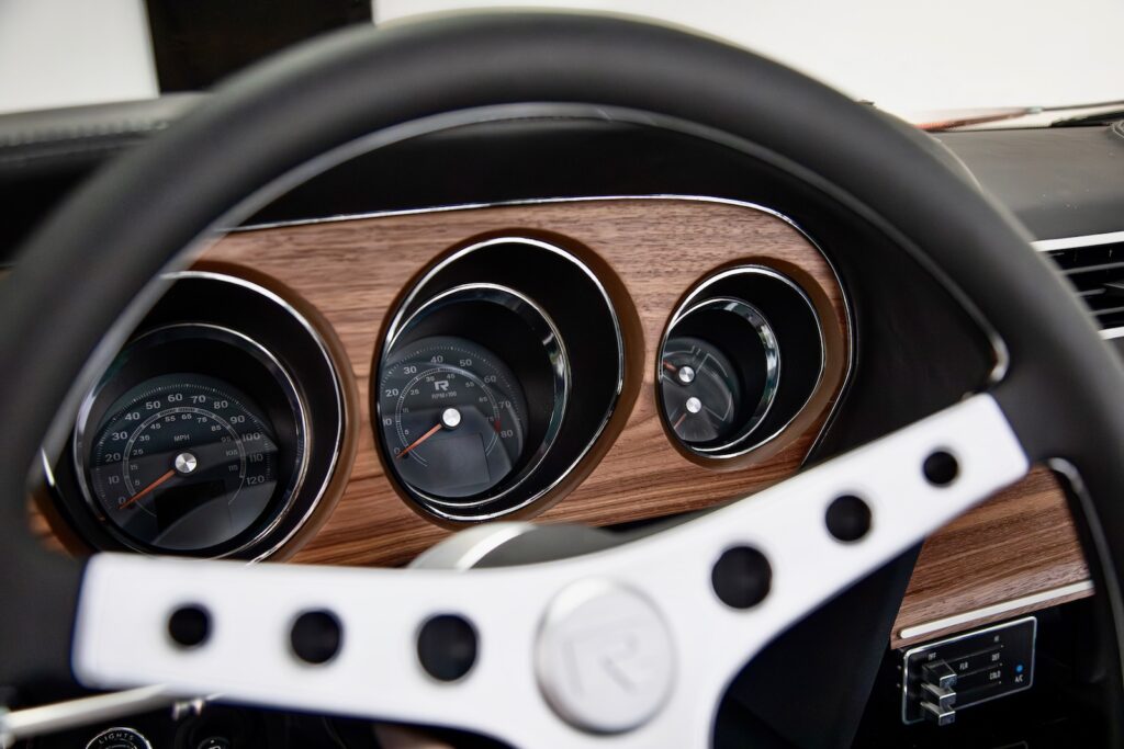 Driver's view of a Revology 1969 Boss Mustang 429 dashboard, featuring a 3-spoke leather-wrapped steering wheel with a silver "R" logo center cap, a walnut wood veneer instrument panel, and modern LED-backlit analog gauges with integrated digital displays.