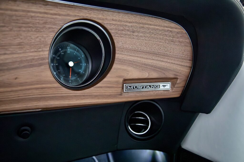 Close-up of the passenger-side dashboard in a Revology 1969 Mustang Boss 429, featuring a genuine walnut wood veneer panel, an analog clock with a red needle, a chrome "MUSTANG" emblem, and a modern circular air conditioning vent.