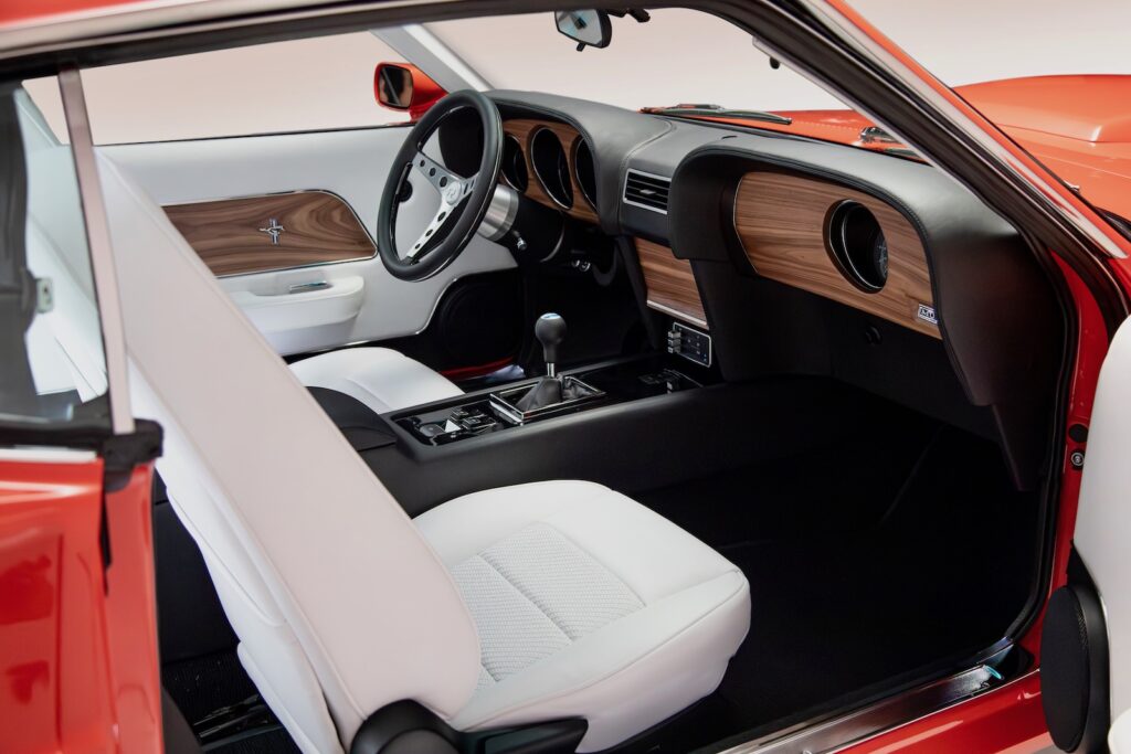 Wide-angle view of the custom interior in a Revology 1969 Ford Mustang Boss 429, featuring parchment white leather bucket seats, a black leather dashboard with walnut wood accents, a 6-speed manual shifter, and a bespoke center console.