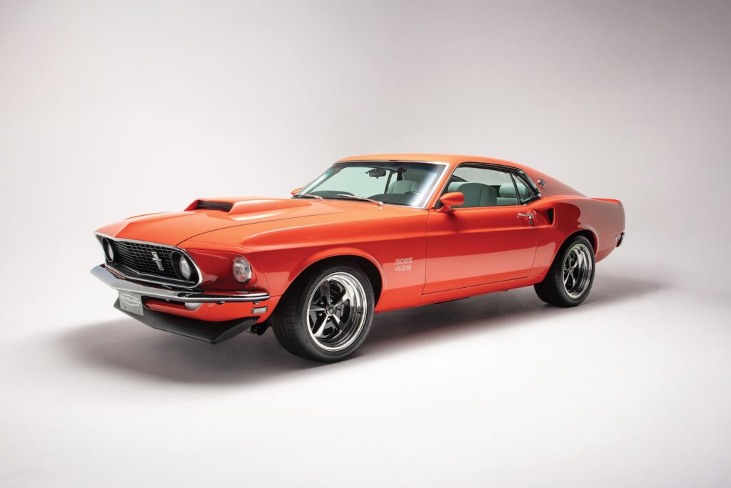 Full front three-quarter studio portrait of a Revology 1969 Ford Mustang Boss 429 restomod in high-gloss orange, showcasing the fastback roofline, functional side scoops, and iconic Boss 429 hood scoop.