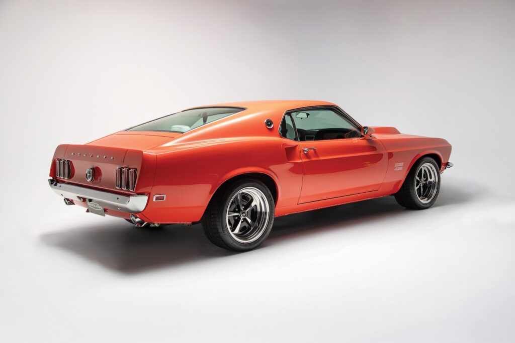 Rear three-quarter studio view of a Revology 1969 Ford Mustang Boss 429 restomod in orange, featuring the classic fastback roofline, chrome rear bumper, tri-bar taillights, and modern wide-tread performance tires.