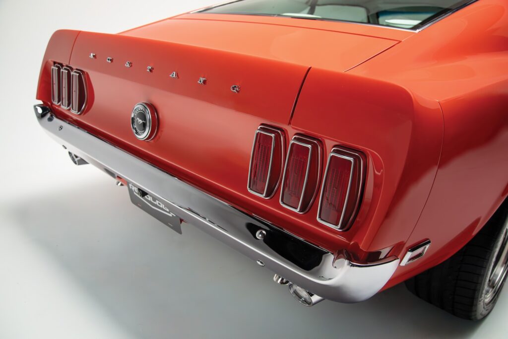 Rear tail panel and tri-bar taillights of an orange Revology 1969 Ford Mustang Boss 429 restomod, featuring individual chrome "MUSTANG" lettering and a high-gloss show-chrome bumper.