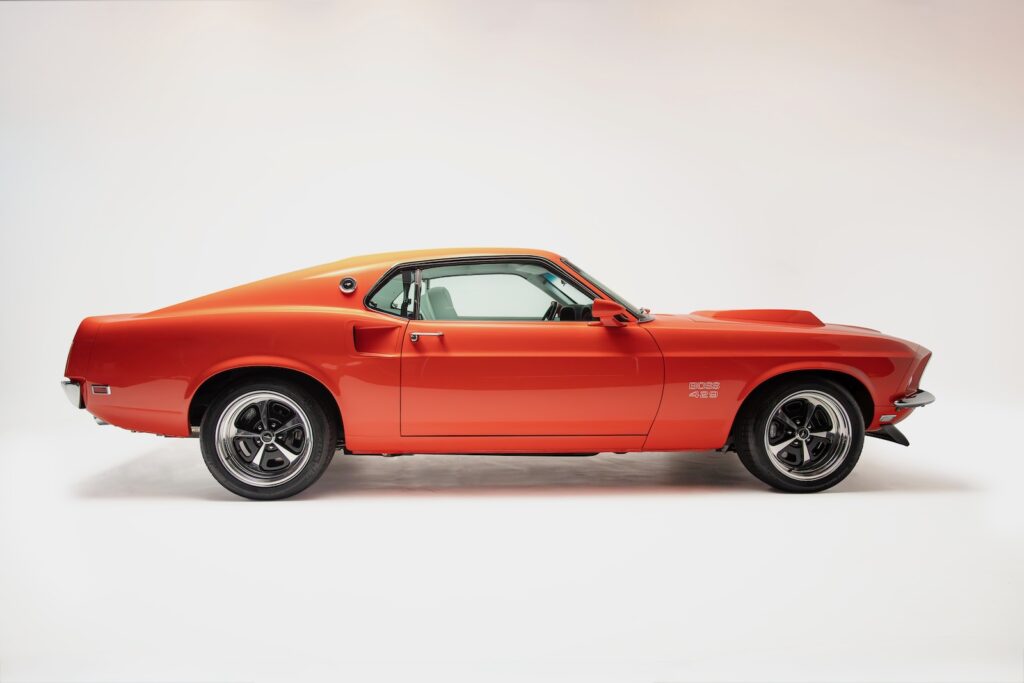 Full side profile of a Revology 1969 Ford Mustang Boss 429 restomod in vibrant orange, featuring a lowered performance stance, modern Magnum 500 wheels, and the iconic Boss 429 hood scoop.