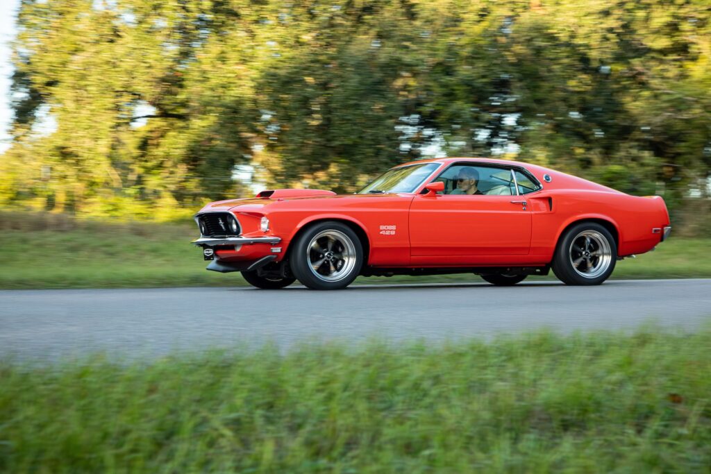 A dynamic motion side profile of a high-performance bright red 1969 Ford Mustang Boss 429 restomod by Revology Cars, accelerating on an open asphalt country road. The photograph uses panning to blur the background trees into streaks, emphasizing speed. The driver is visible inside. The visible fender features the 'Boss 429' script, and the modern 17-inch Magnum 500-style wheels and high-gloss paint are prominent.
