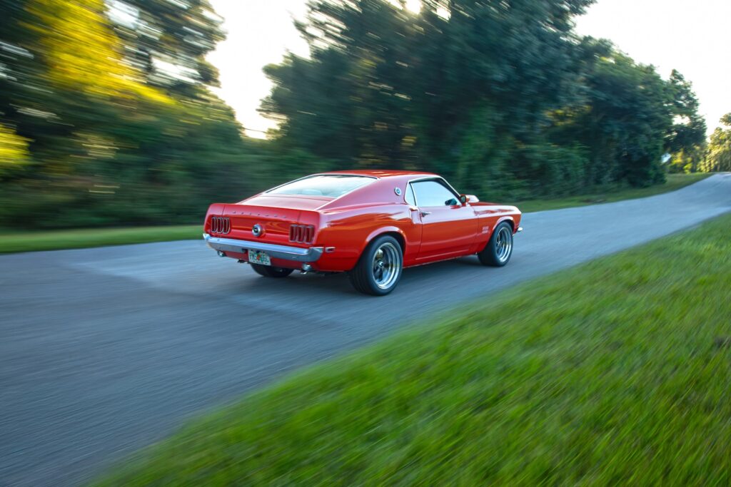 Rear three-quarter motion shot of an orange Revology 1969 Ford Mustang Boss 429 restomod accelerating on a country road, highlighting the fastback silhouette and modern performance stance.