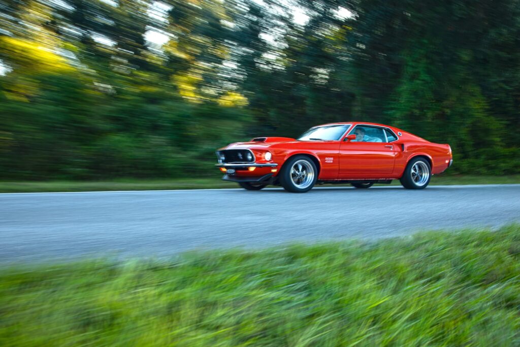 Front three-quarter motion shot of a vibrant orange Revology 1969 Ford Mustang Boss 429 restomod accelerating on a paved road, highlighting the iconic quad-headlights and aggressive Boss 429 hood scoop.