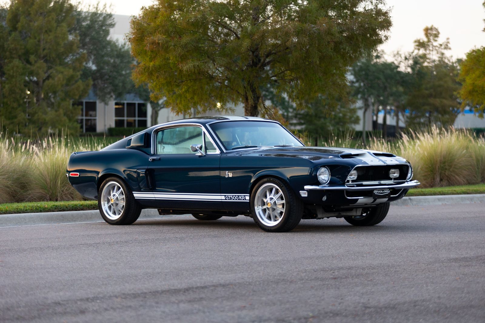 Front-quarter view of a dark green 1968 Revology Shelby GT500KR Fastback with white rocker stripes and polished multi-spoke wheels, parked on a road.