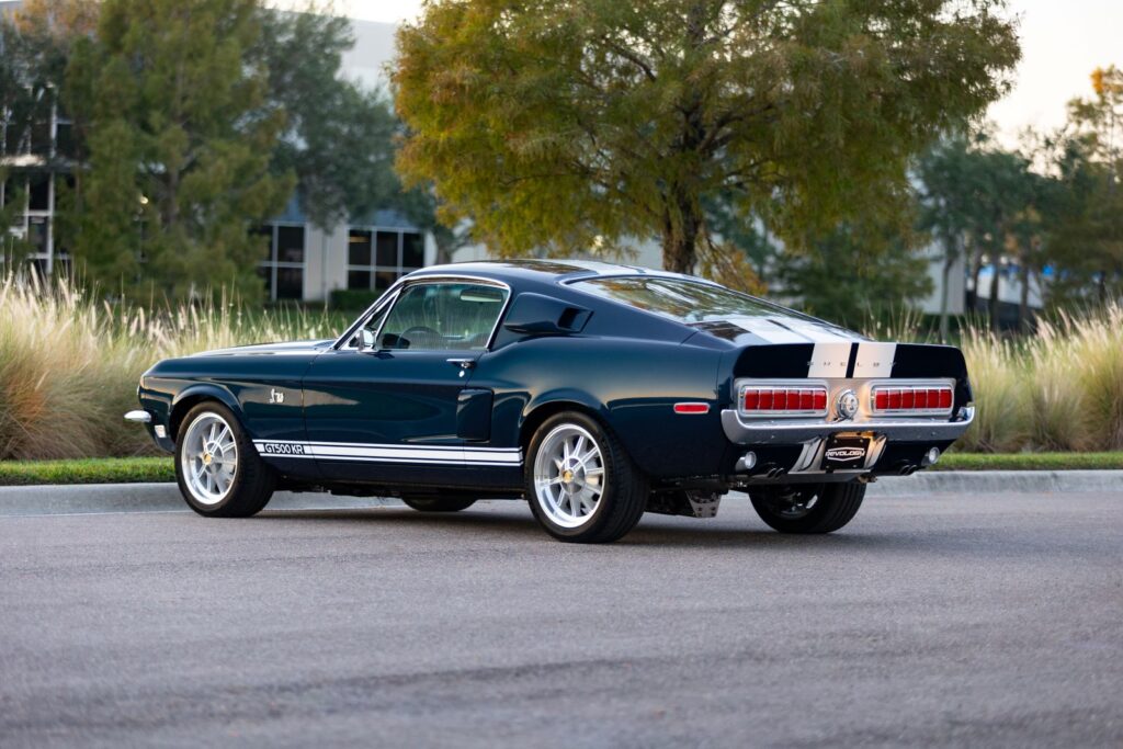 Rear-quarter view of a dark green 1968 Revology Shelby GT500KR Fastback featuring iconic horizontal sequential taillights and white Le Mans racing stripes.