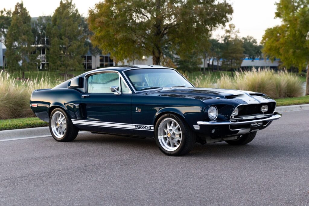 Front three-quarter view of a 1968 Revology Shelby GT500KR in dark green, showcasing the unique 1968 hood scoops and white G.T.500KR side stripes.