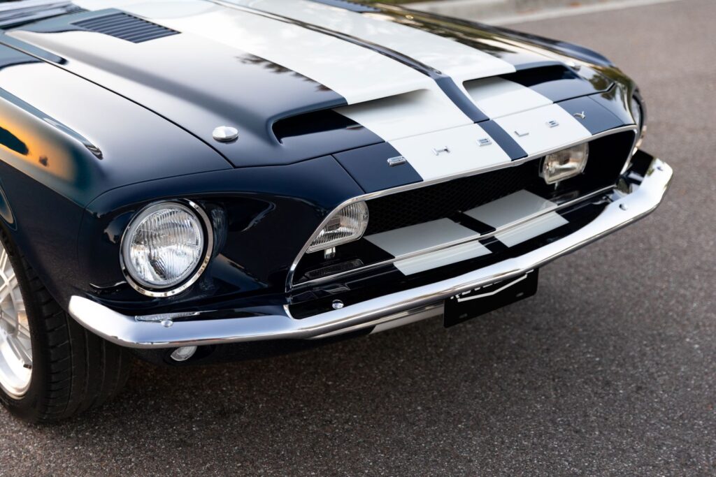 Close-up of the front nose and hood of a 1968 Revology Shelby GT500KR, showing the functional ram-air scoops, white racing stripes, and Shelby lettering on the header panel.