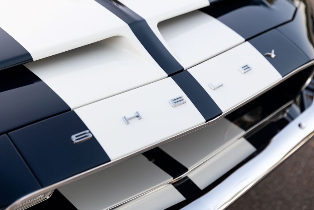 Macro view of the silver "S-H-E-L-B-Y" individual lettering on the front header panel of a 1968 Revology GT500KR, with white Le Mans stripes and functional hood scoops.