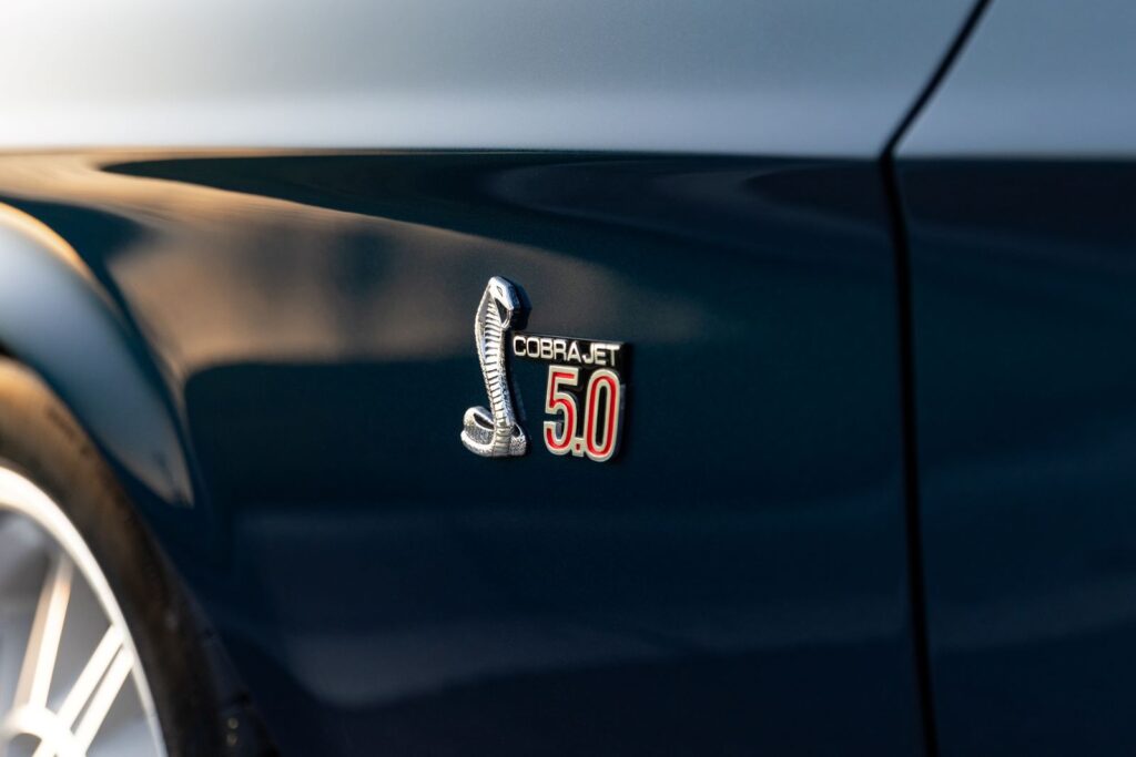 Close-up of a custom "Cobra Jet 5.0" fender emblem on a dark green 1968 Revology Shelby GT500KR, featuring a polished chrome snake and red 5.0 lettering.
