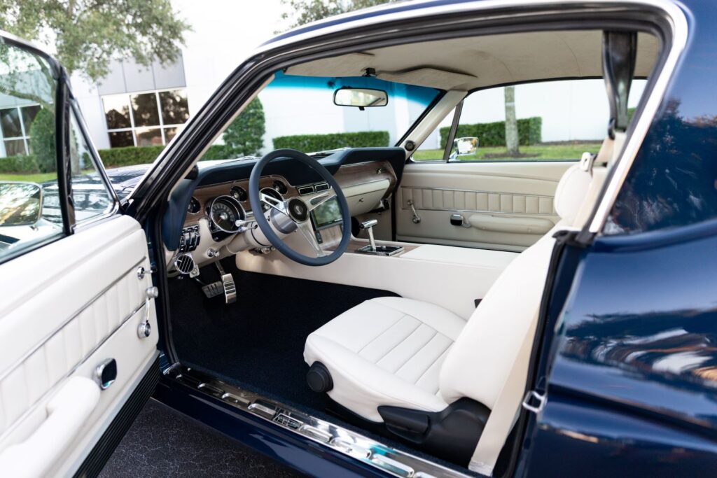 Driver-side view of the white leather interior in a 1968 Revology Shelby GT500KR, featuring a full center console, custom steering wheel, and premium high-back bucket seats.