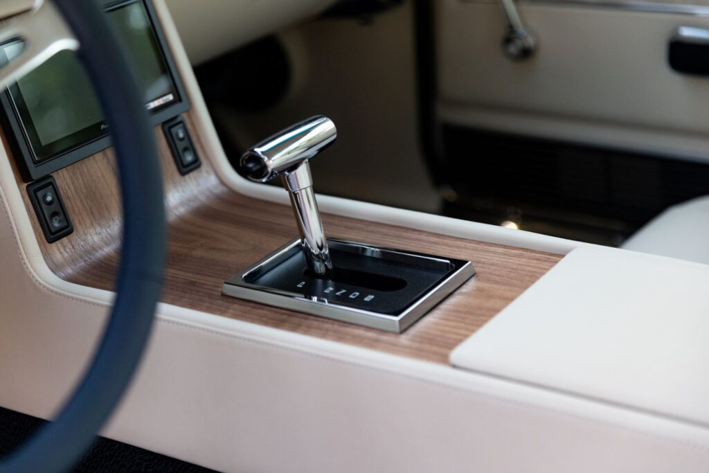 Close-up of a polished chrome automatic gear shifter and wood grain center console in a 1968 Revology Shelby GT500KR with white leather upholstery.