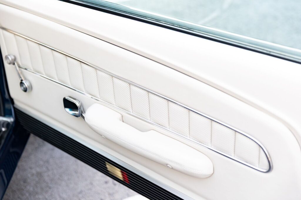 High-angle view of the driver-side door panel in a 1968 Revology Shelby GT500KR, featuring white leather, vertical pleating, and chrome "eyebrow" trim.