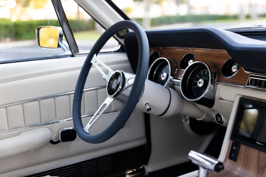 Driver-side perspective of the 1968 Revology Shelby GT500KR interior, showing the leather-wrapped steering wheel, wood grain dash, and white leather door panel.