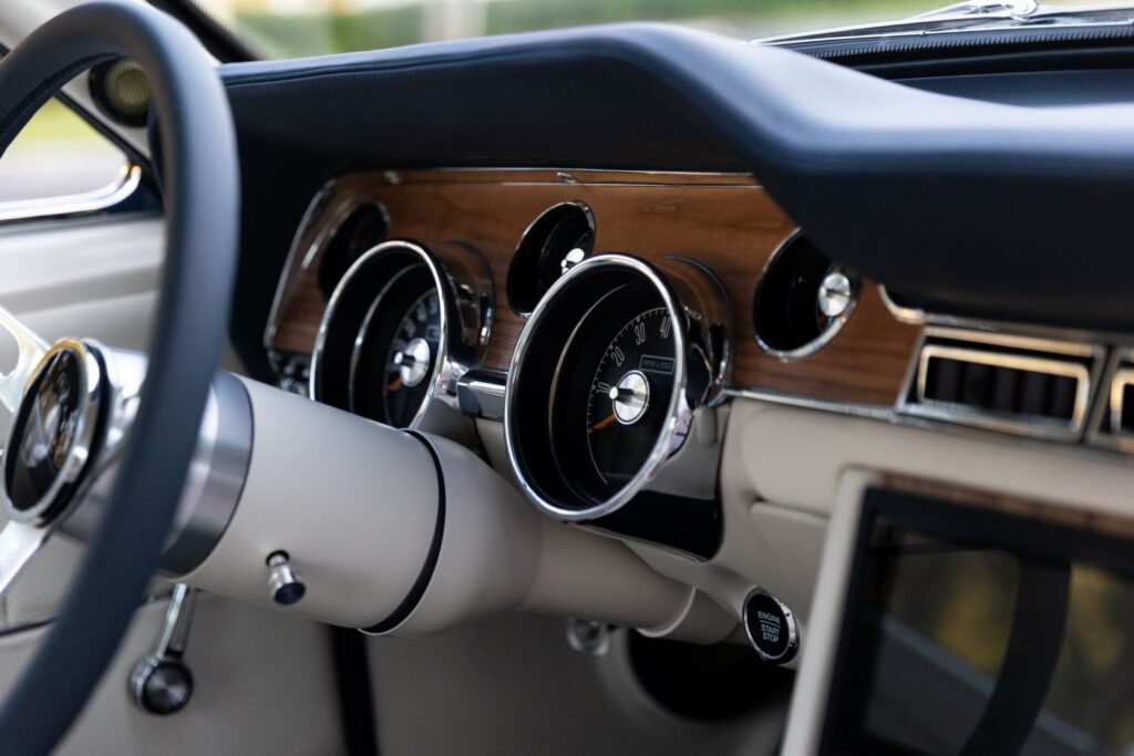 Close-up of the wood grain dashboard and white steering column in a 1968 Revology Shelby GT500KR, featuring a modern "Engine Start" button and chrome-rimmed gauges.