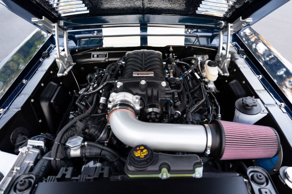 Engine bay of a 1968 Revology Shelby GT500KR featuring a supercharged 5.0L Coyote V8 engine with a custom cold air intake and black blower.